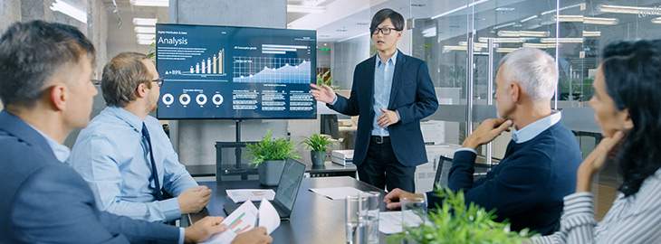 Businessman Gives Report/ Presentation to His Business Colleagues, Pointing at the Results Showing Statistics, Pie Charts and Company's Growth On Wall TV Screen