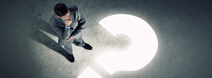businessman standing in front of a glowing question mark projected on the floor in a dark room