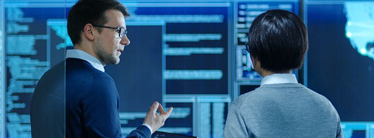 In the System Control Room IT Specialist and Project Engineer Have Discussion while Holding Laptop, they're surrounded by Multiple Monitors with Graphics.