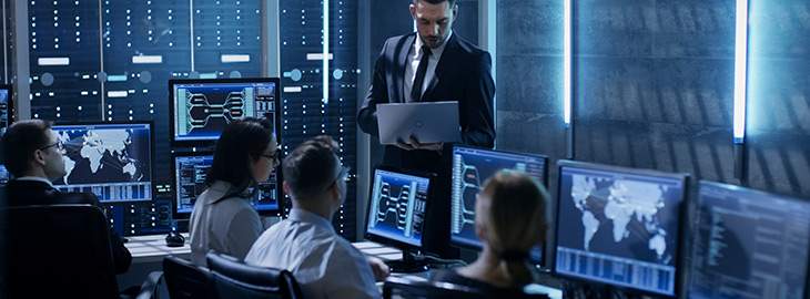 Professional IT Engineers Working in System Control Center Full of Monitors and Servers. Supervisor Holds Laptop and Holds a Briefing