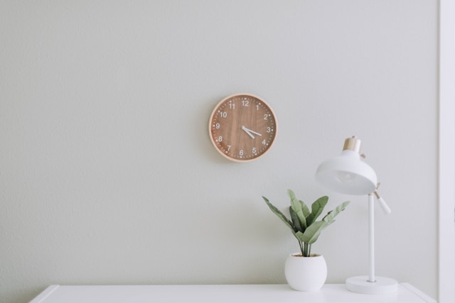 wooden clock on white wall, above white desk with lamp and plant