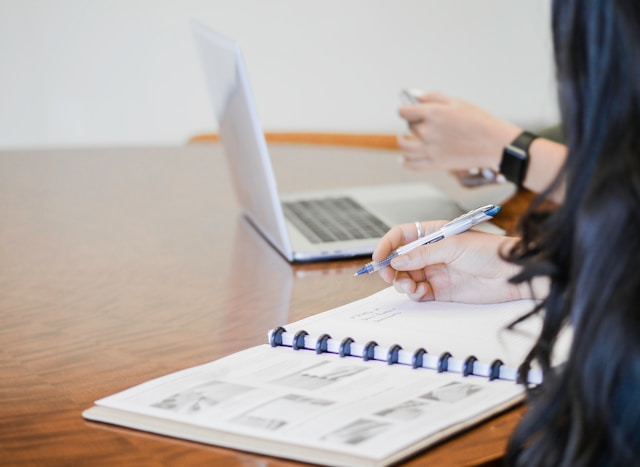 two people working at wooden desk, with notebook and laptop on table